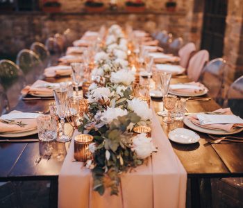 in backyard of villa in Tuscany there is banquet wooden table decorated with cotton and eucalyptus compositions, glasses, burning candles and plates are placed on table, evening